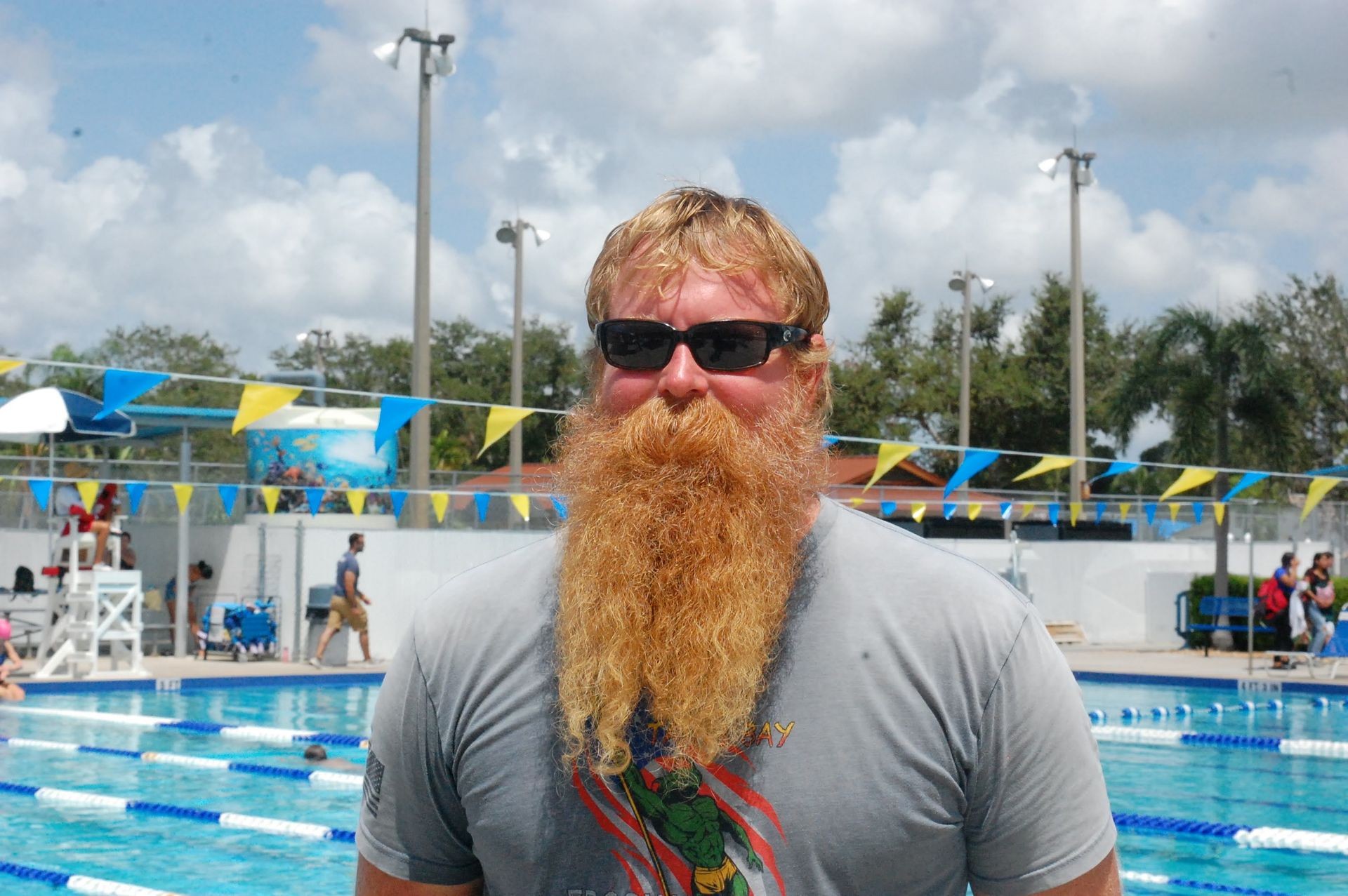 Man with long red beard and sunglasses at a poolside, wearing a gray T-shirt.