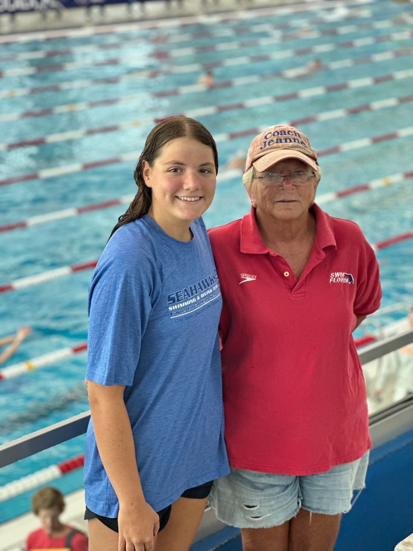 Two women smiling in front of an indoor swimming pool with lanes.