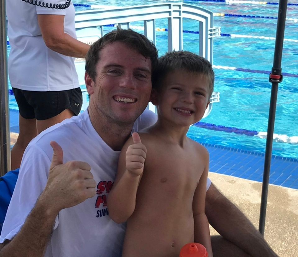 Man and child at a swimming pool giving thumbs up, with another person standing in the background.