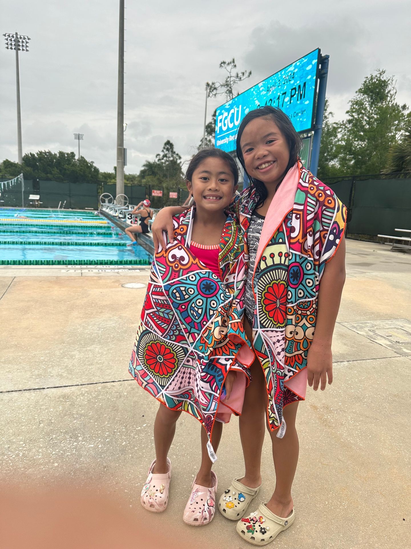 Two kids smiling, wearing colorful towels near a swimming pool with a digital scoreboard in the background.